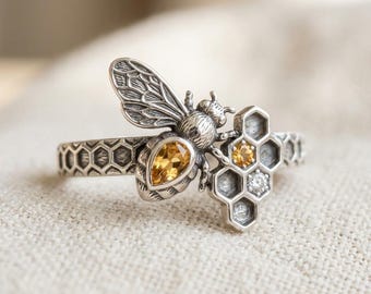 May include: A silver ring with a bee motif, featuring a pear-cut yellow gemstone and a honeycomb design. The ring incorporates a cluster of hexagonal shapes with yellow and clear stones, showcasing intricate detailing.