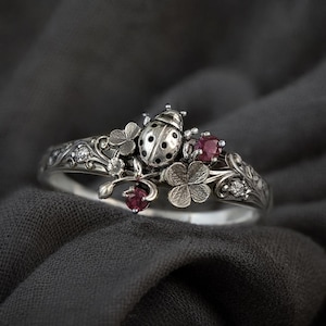May include: A silver ring featuring a ladybug, clovers, and small red gemstones. The ring has intricate floral detailing and small clear gemstones. The ladybug has black spots and is centered on the ring.