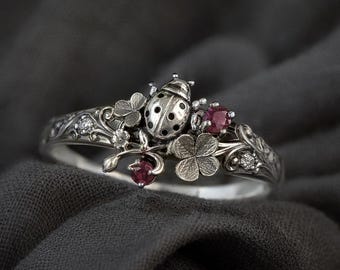 May include: A silver ring featuring a ladybug, clovers, and small red gemstones. The ring has intricate floral detailing and small clear gemstones. The ladybug has black spots and is centered on the ring.