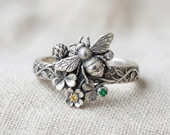 May include: A sterling silver ring featuring a detailed bee centrepiece surrounded by sculpted clover leaves and honeycomb patterns. The band includes a small yellow gemstone, a green gemstone, and a clear stone set among the metal floral elements.