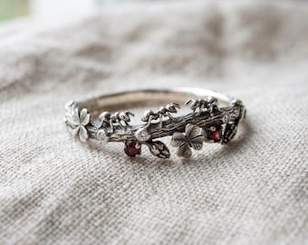May include: A silver ring designed to resemble a textured tree branch, featuring sculpted ants crawling along the surface, small four-leaf clover accents, embedded clear stones, and two round, deep red garnet gemstones set into the metal band.