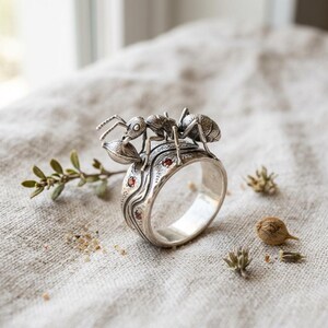 Può includere: Un anello in argento con una formica scolpita che trasporta una piccola foglia sul dorso. La fascia presenta un design testurizzato e ondulato con tre piccole pietre preziose rotonde di colore bruno-rossastro incastonate sul lato.