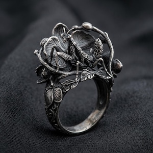 May include: A silver ring featuring a detailed praying mantis perched atop a cluster of sculpted flowers and leaves. The metal has an oxidised finish that highlights the intricate textures and patterns on the insect and the surrounding botanical elements.