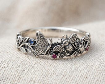 May include: A silver ring featuring a detailed relief of a butterfly, a bee, and a small beetle amidst floral vines and honeycomb patterns. The band is accented with small, round, faceted blue, white, and red gemstones set within the metal design.