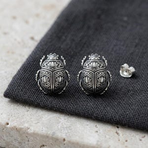 May include: A pair of silver-toned stud earrings shaped like scarab beetles. Each earring features intricate engraved details, including a lotus flower motif on the wing covers and geometric patterns along the edges, accompanied by a metal earring back.