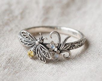 May include: A silver ring featuring a detailed insect with a yellow gemstone on its body, positioned next to two small flowers with iridescent stones. The band is decorated with an intricate engraved scrollwork pattern throughout its circumference.