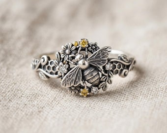 May include: A silver ring featuring a central bee motif surrounded by small flowers and leaves. The band incorporates a honeycomb pattern on the sides. The design is accented with small, clear white stones and tiny yellow stones embedded within the floral elements.