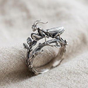 Op de afbeelding: Een zilveren ring met een gedetailleerde bidsprinkhaan op een takontwerp. De ringband is vormgegeven als een knoestige twijg met kleine blaadjes en knoppen, uitgevoerd in gepolijst zilver met donkere accenten in de dieper gelegen delen.