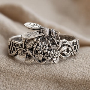 Pode incluir: Uma pulseira de prata com uma abelha detalhada pousada sobre um grupo de flores e folhas. A banda é decorada com um padrão hexagonal de favo de mel em ambos os lados, trabalhada com um acabamento oxidado para realçar as texturas.