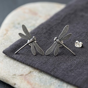 May include: A pair of silver-toned dragonfly stud earrings featuring intricate wing detailing and textured bodies. Each earring has a post back and is accompanied by a separate silver-toned butterfly clutch earring back for secure fastening.