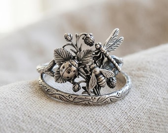 May include: A silver ring featuring a detailed nature-inspired design with a ladybird, a bee, and a dragonfly perched among leaves and small berries. The band of the ring has an embossed vine pattern, and the entire piece has an oxidised finish.