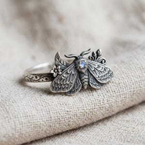 May include: A silver ring featuring a detailed moth design with textured wings, a small round iridescent stone set in the center of the moth, and a band decorated with engraved star and vine patterns along the outer surface of the metal band.