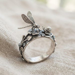 May include: A silver ring featuring a detailed dragonfly and a small snail perched on top of sculpted leaves. The band is decorated with embossed plant motifs and small, light blue gemstones embedded along the sides of the metal surface.