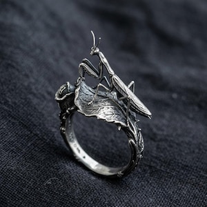 Op de afbeelding: Een zilveren ring met een gedetailleerde bidsprinkhaan die op een gestructureerd blad zit. De ringband is ontworpen met complexe botanische elementen, waaronder een kleine rozenknop, en heeft een geoxideerde afwerking voor extra detail.
