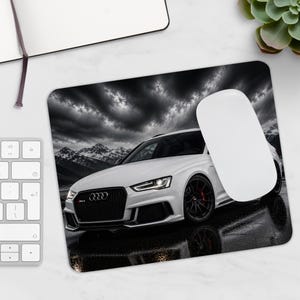 May include: A rectangular mouse pad featuring a high-contrast image of a white Audi RS4 parked on a wet surface. The car is positioned against a backdrop of dark, dramatic clouds and snow-capped mountains, with a white computer mouse resting on top.