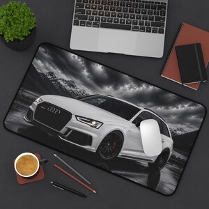 May include: A rectangular desk mat featuring a black and white photograph of a white Audi RS6 station wagon parked against a backdrop of mountains and a cloudy sky. The mat is positioned on a dark surface with a laptop, notebook, and coffee cup.