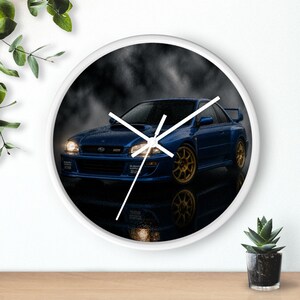 May include: A round wall clock featuring a blue Subaru Impreza 22B car with gold wheels on the face. The car is depicted against a dark, cloudy background with its headlights illuminated. The clock has a white frame and simple white hands.