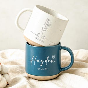 Peut inclure: Une pile de deux tasses en céramique. La tasse supérieure est blanc cassé avec des taches, un dessin floral, les lettres h + q et la date 09.23.2026. La tasse inférieure est bleue avec le nom Hayden, un petit cœur et la date 08.26.26.