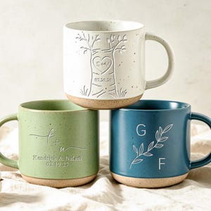 May include: Three ceramic mugs with speckled finishes and unglazed bases. The top white mug features an embossed tree with a heart containing S+V and the date 07.20.26. The green mug shows K+A with Kendrick & Naladi 02.19.27. The blue mug displays G and F.