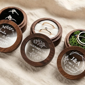 Puede incluir: Cuatro cajas redondas de madera para anillos con tapas transparentes que presentan texto grabado en blanco y diseños florales. Las cajas contienen anillos sobre bases de terciopelo negro, blanco o musgo verde. El texto incluye nombres y fechas.