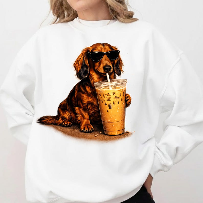 Peut inclure: Sweat-shirt blanc avec un dessin d'un teckel marron portant des lunettes de soleil et buvant du caf&eacute; glac&eacute; dans un gobelet transparent. Le chien est assis sur du sable. Design graphique amusant et d&eacute;contract&eacute;.