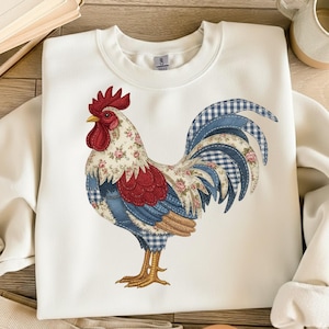 May include: White sweatshirt featuring a colorful rooster design. The rooster is made of patchwork fabric with floral, gingham, and solid blue patterns. The rooster has a red comb and wattle, and yellow feet. The sweatshirt is a Gildan brand.