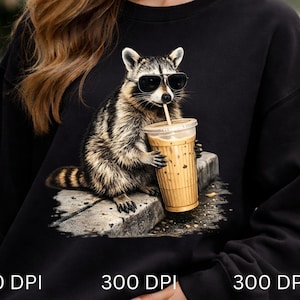 May include: Black sweatshirt featuring a raccoon wearing sunglasses and drinking from a coffee cup with a straw. The raccoon is depicted in a realistic style, with detailed fur and a relaxed pose. The sweatshirt is a solid black color.
