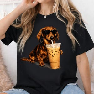 Peut inclure: T-shirt noir avec un dessin d'un teckel marron portant des lunettes de soleil et tenant un caf&eacute; glac&eacute;. Le chien a des poils d&eacute;taill&eacute;s et est dans une pose d&eacute;tendue, avec la tasse de caf&eacute; dans sa patte.