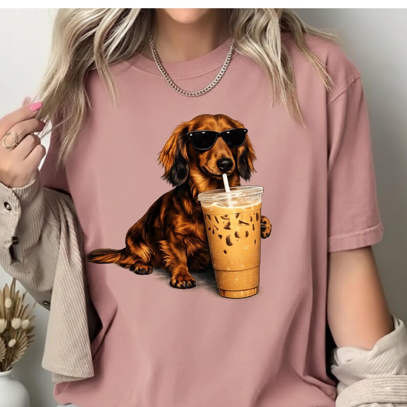 Peut inclure: T-shirt rose poudr&eacute; avec un motif de teckel portant des lunettes de soleil et buvant du caf&eacute; glac&eacute;. Le chien est marron et noir, et le caf&eacute; est dans un gobelet en plastique transparent avec une paille.