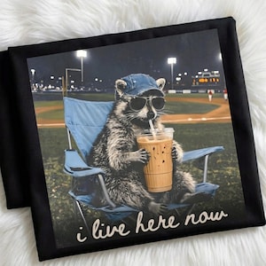 May include: Black t-shirt with a graphic of a raccoon wearing sunglasses and a baseball cap, drinking iced coffee while seated in a blue chair. The text "i live here now" is written in white script below the image.