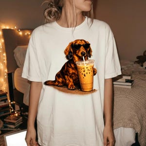 Peut inclure: T-shirt blanc avec un motif d'un teckel marron portant des lunettes de soleil et buvant du caf&eacute; glac&eacute;. Le chien est repr&eacute;sent&eacute; avec des d&eacute;tails de fourrure et tient un gobelet en plastique transparent avec une paille. Le t-shirt est ample.