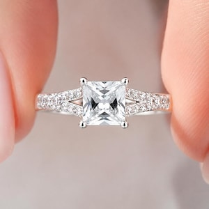 May include: A silver-toned ring featuring a large, square-cut, clear centre stone held by four prongs. The band is split into two sections near the centre stone, with both sides encrusted with small, round, clear stones along the shank.