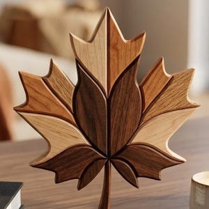 May include: A wooden maple leaf sculpture with a multi-toned design. The leaf is crafted from various shades of brown wood, creating a layered effect. The sculpture is displayed on a wooden surface, showcasing the natural wood grain.