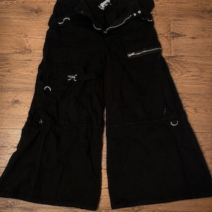 May include: Black wide-leg pants featuring multiple utility pockets, silver-toned metal hardware, including D-rings and zippers, and a button-fly closure. The garment has a loose, flared silhouette with horizontal seam detailing across the legs.
