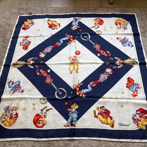 May include: A square silk scarf featuring a dark blue border and a central diamond pattern on a cream background. The design displays multiple colorful illustrations of clowns performing circus acts, including juggling and unicycling, with the Chopard logo.