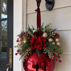 May include: A large, shiny red spherical bauble hanging from a dark metal wall hook by a deep red velvet ribbon. The top is decorated with green pine needles, red berries, small gold glittery spheres, and a central red poinsettia flower.