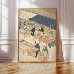 May include: A framed print of a Japanese woodblock print depicting a pottery market scene. The artwork shows people working at stalls with pottery, under a blue and beige awning. The print has a beige background and is framed in a gold-colored frame.