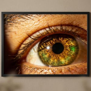 Puede incluir: Impresión enmarcada de primer plano de un ojo humano. El iris es de un llamativo color marrón dorado con reflejos verdes y amarillos. Largas pestañas enmarcan el ojo, y la piel alrededor del ojo es de un marrón cálido y claro.