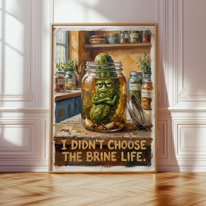 May include: A framed art print featuring a grumpy pickle character inside a jar, with the text "I didn't choose the brine life." The artwork has a rustic, cartoonish style, with a kitchen setting in the background.