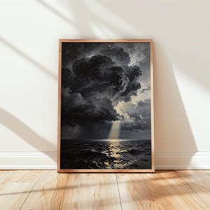 Puede incluir: Una obra de arte enmarcada representa un dramático paisaje marino bajo un cielo tormentoso. La pintura presenta nubes oscuras y ondulantes, un mar turbulento y un rayo de sol que se abre paso. El marco de madera añade un toque clásico.