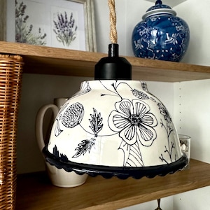 Può includere: Una lampada a sospensione in ceramica bianca con un motivo floreale nero e un bordo smerlato blu navy. La lampada è appesa a un cavo di corda intrecciata. La lampada si trova in una stanza con mensole in legno e oggetti decorativi.