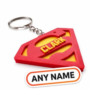 以下が含まれることがあります： 赤と黄色のスーパーマンのロゴキーホルダー。黄色の文字で「CLARK」と書かれています。キーホルダーには、シルバーのキーリングと、黒い文字で「ANY NAME」と書かれた白いステッカーが付いています。
