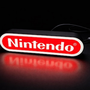 May include: A rectangular black light-up sign with the Nintendo logo in red and white. The sign is illuminated, casting a red reflection on the black surface below. A power cord extends from the back.