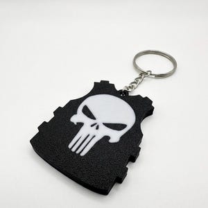 May include: Black and white keychain featuring a skull design on a black vest-shaped pendant. The keychain has a silver ring and chain. The skull is white with black eye sockets and a nose cavity. The vest has a textured surface.