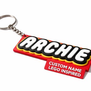 Può includere: Un portachiavi rosso, giallo e nero con il nome "ARCHIE" a lettere bianche. Il portachiavi ha anche un piccolo tag rosso con la scritta "CUSTOM NAME LEGO INSPIRED". Il portachiavi è attaccato a un portachiavi argentato.