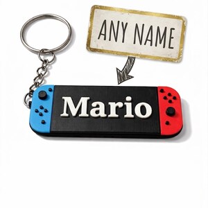 以下が含まれることがあります： ゲームコントローラーのようにデザインされた、白で「Mario」という名前が入った黒いキーホルダー。 コントローラーには青と赤のアクセントがあります。 上のタグには「ANY NAME」と書かれています。