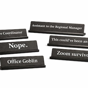 Könnte beinhalten: Eine Sammlung schwarzer Schreibtisch-Namensschilder mit weißem Text. Die Namensschilder lauten: "Chaos Coordinator", "Nope", "Office Goblin", "Assistant to the Regional Manager", "This could've been an email" und "Zoom survivor."