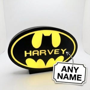 May include: A black and yellow Batman logo with the name "HARVEY" in white. The logo is on a black base. A white tag with the words "ANY NAME" is attached to the logo.