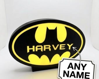 May include: A black and yellow Batman logo with the name "HARVEY" in white. The logo is on a black base. A white tag with the words "ANY NAME" is attached to the logo.
