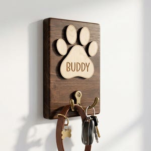 Pode incluir: Um porta-chaves de madeira castanha escura com um design de pegada de pata e o nome "BUDDY" gravado. Possui dois ganchos de latão e segura uma coleira de cão e chaves.